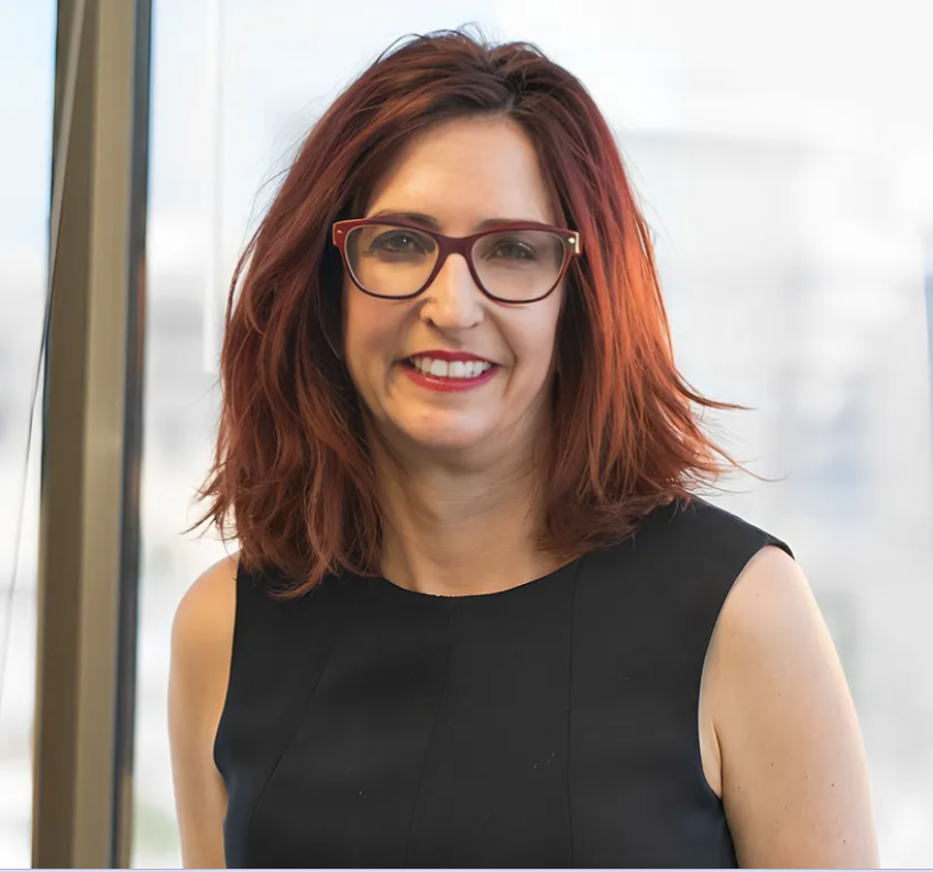 A woman with red hair and glasses is smiling.