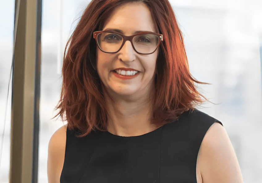 A woman with red hair and glasses is smiling.